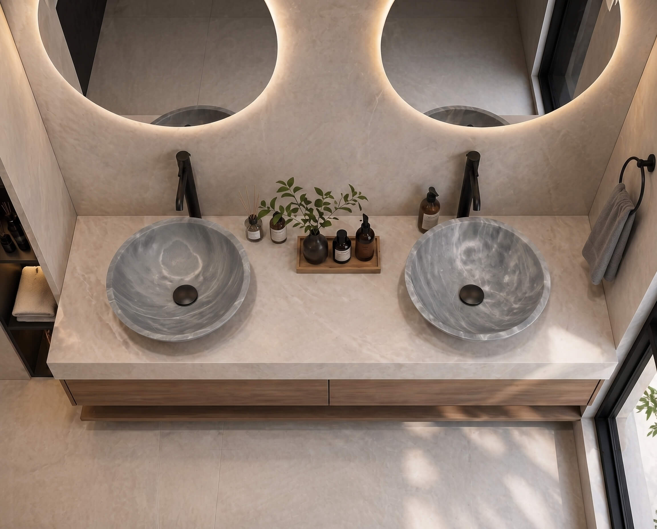 Two Carrara gray marble round vessel sinks on a wall-mounted walnut vanity against a large-format limestone wall in a Japandi-style bathroom, matte black wall-mounted curved faucets, dual round halo-lit mirrors, wooden tray with botanical accents and amber apothecary bottles.