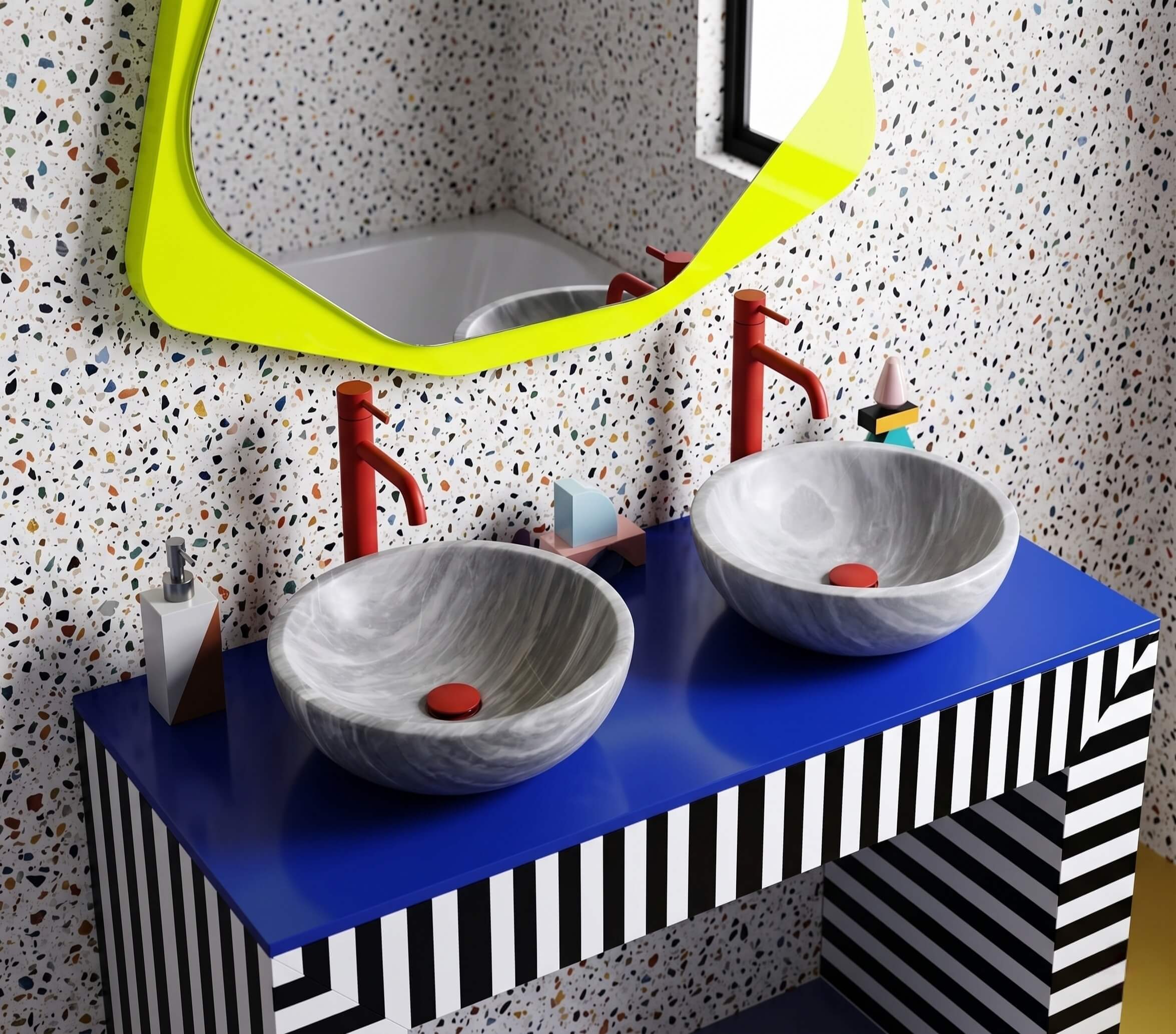 Two Carrara gray marble round vessel sinks serving as neutral focal points on a cobalt blue countertop atop a black-and-white striped Memphis-style vanity, vermillion red gooseneck faucets and matching drain caps, multi-color terrazzo wall, neon yellow asymmetric mirror, demonstrating natural marble compatibility with bold maximalist palettes.