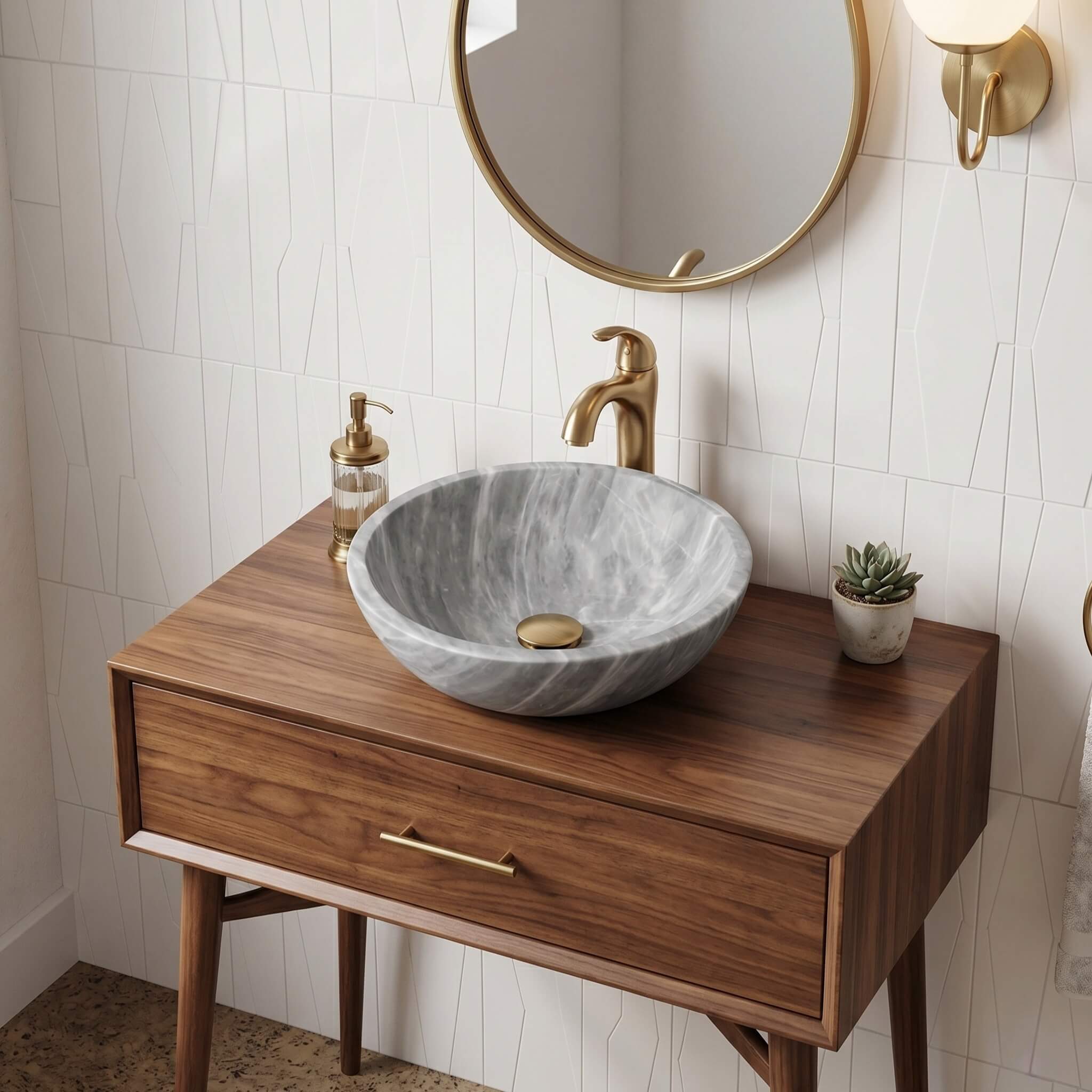 Carrara gray marble round vessel sink atop a mid-century walnut vanity with tapered legs and brass drawer pull, brushed brass tall faucet and round mirror, white 3D geometric ceramic tile backsplash, cork tile flooring, succulent accent.