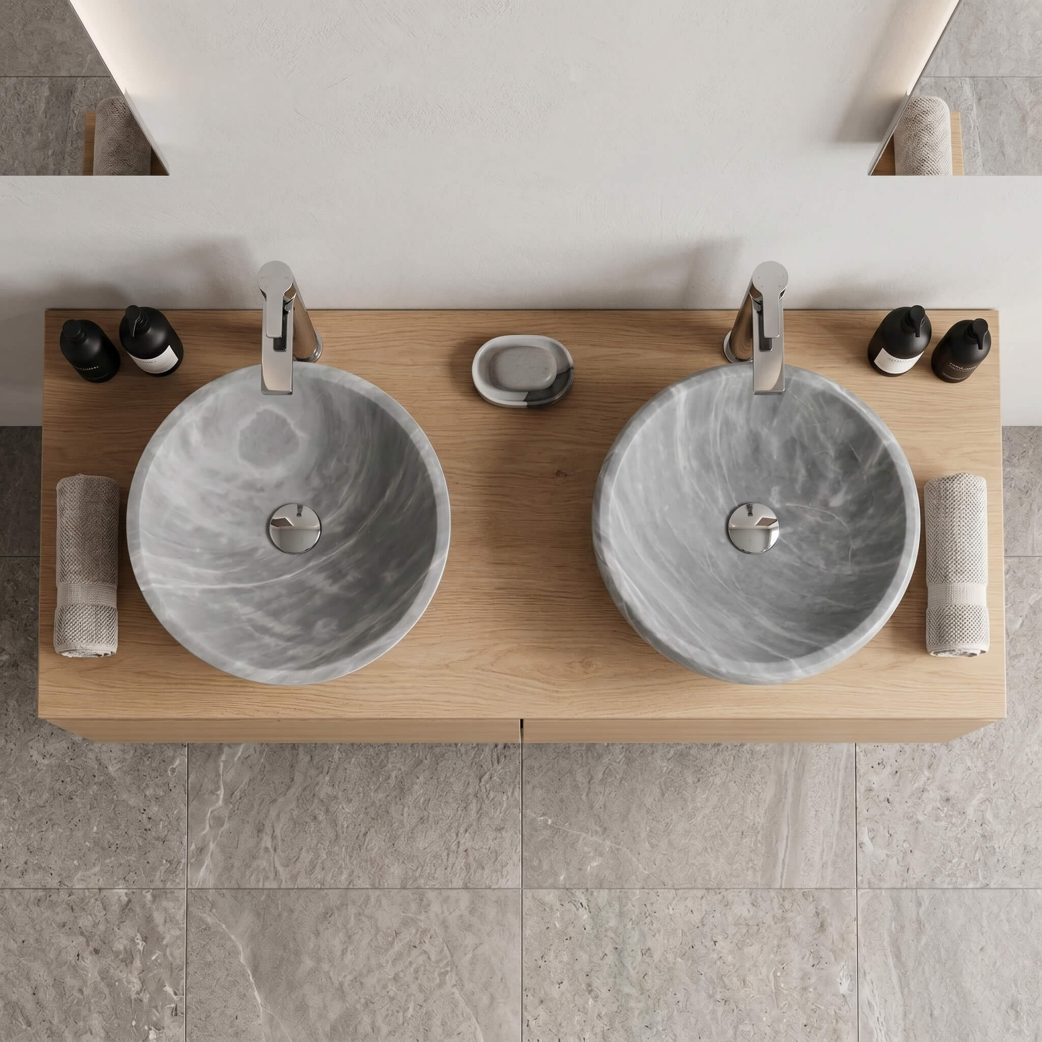 Overhead top-down view of two Carrara gray marble round vessel sinks on a light oak wood vanity top in a minimalist bathroom, chrome vessel faucets, rolled linen towels, matte black accessories, gray stone tile floor visible below.