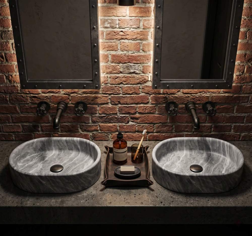 Pair of oval Carrara gray marble vessel sinks on concrete countertop against exposed red brick wall with black pipe faucets.
