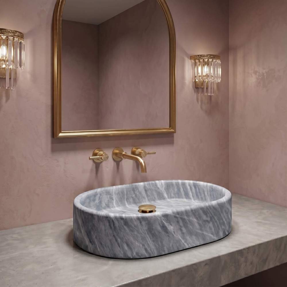 Oval Carrara gray marble vessel sink on travertine counter with pink plaster wall, brushed gold knurled faucet, arched mirror.