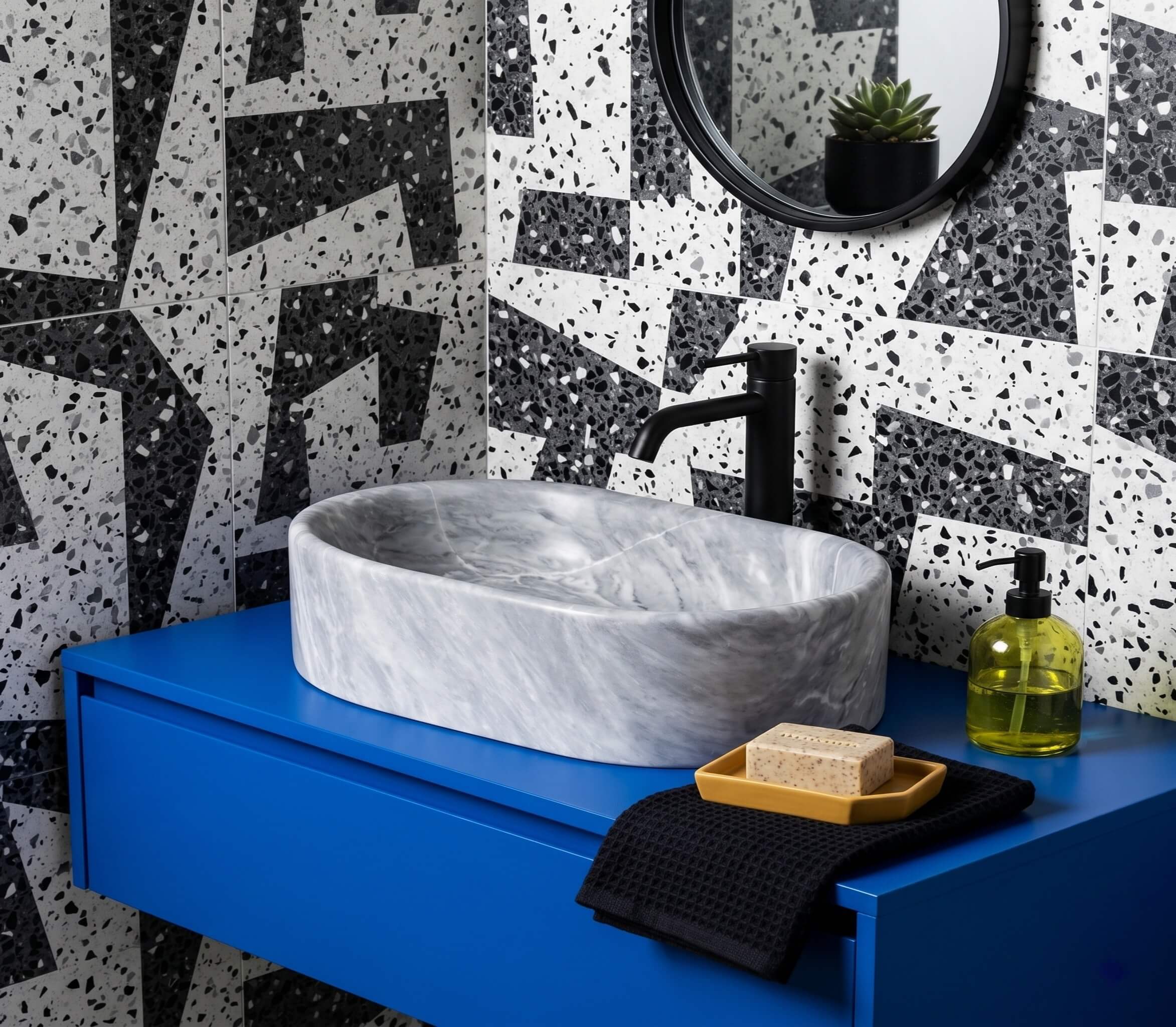 Oval Carrara gray marble vessel sink on cobalt blue floating vanity against black-white terrazzo tile with matte black faucet.