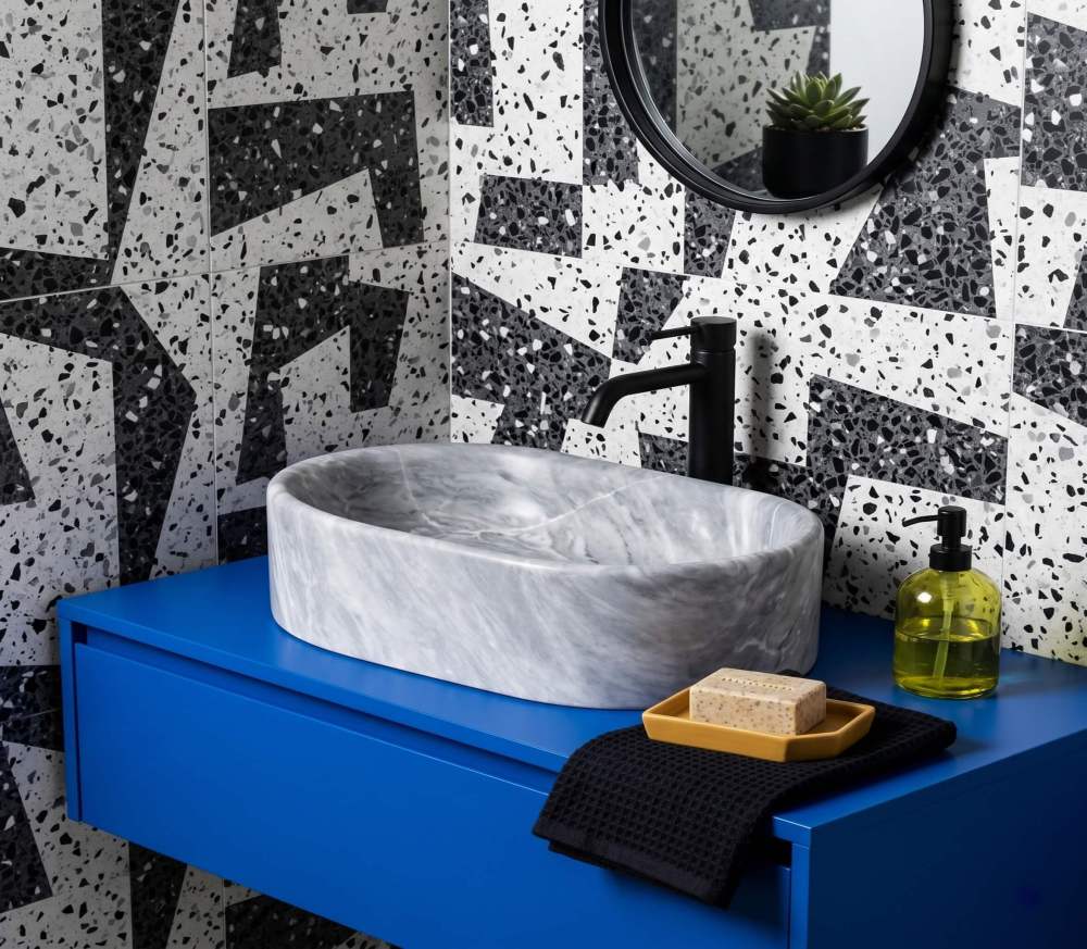 Oval Carrara gray marble vessel sink on cobalt blue floating vanity against black-white terrazzo tile with matte black faucet.