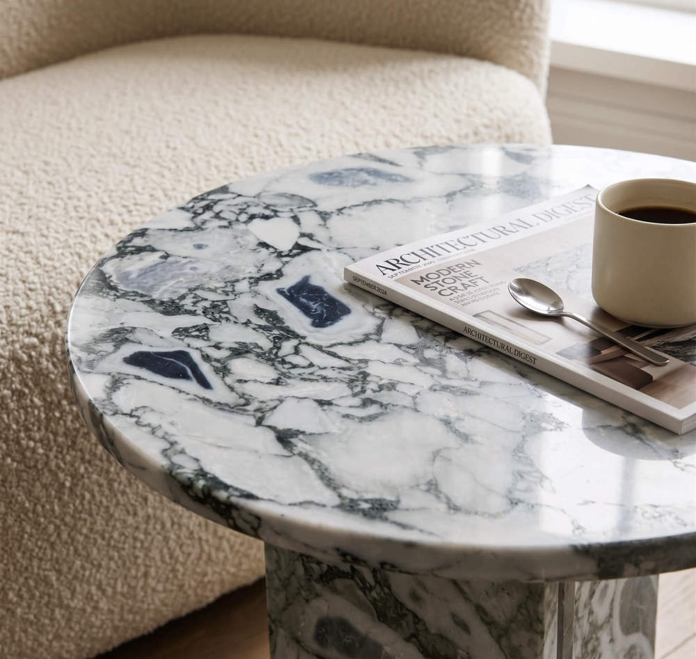 Close-up detail of cloudy blue marble side table showing polished beveled edge, swirling veining pattern, and Architectural Digest magazine.
