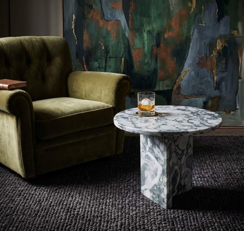 Cloudy blue marble side table in moody whisky lounge with olive tufted velvet armchair, abstract painting, and crystal glass with ice sphere.