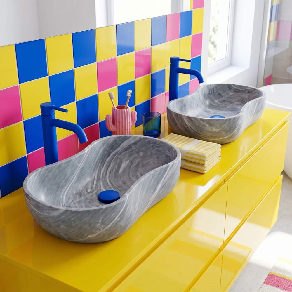 Two Carrara gray marble vessel sinks on glossy yellow vanity with cobalt blue faucets and color-block tile wall in a maximalist pop-art bathroom.
