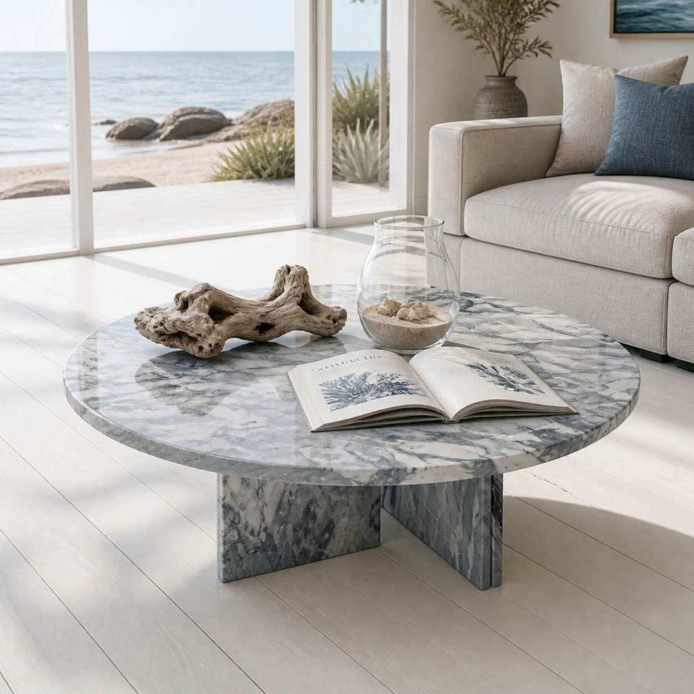 Cloudy blue marble coffee table in coastal beach house living room with driftwood, shell jar, linen sofa, and ocean view beyond.