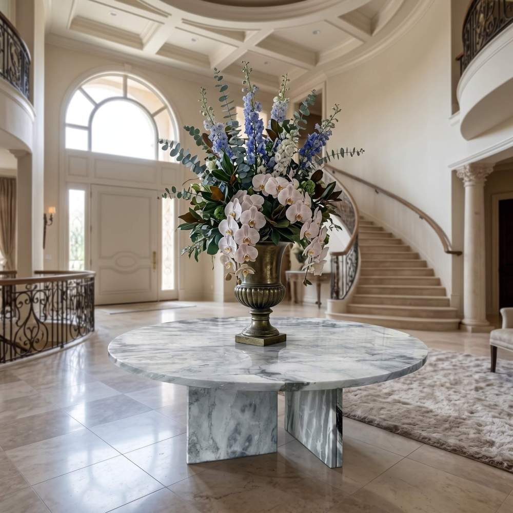 Cloudy blue marble round table as grand mansion foyer centerpiece with bronze urn, orchid arrangement, and sweeping staircase backdrop.