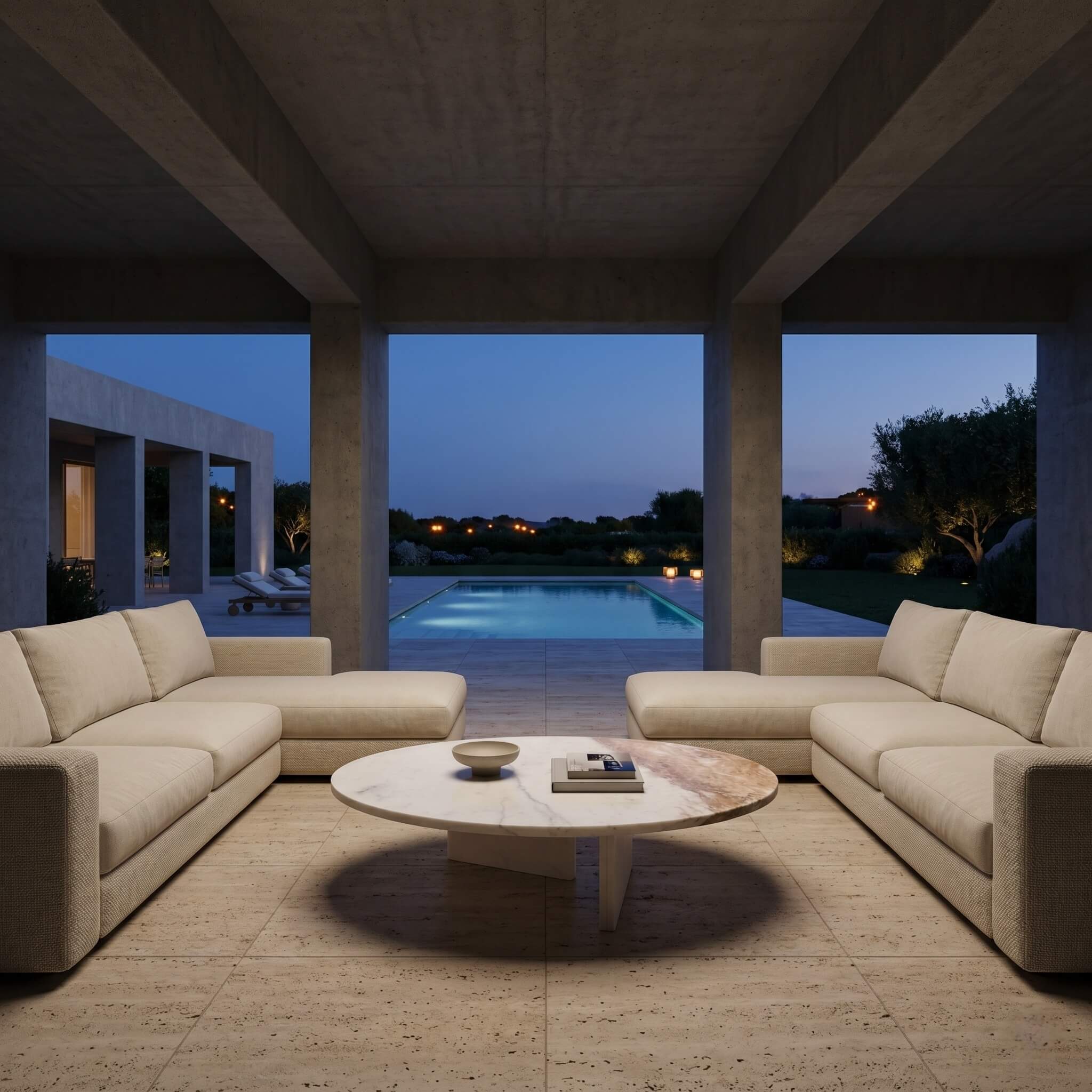 Round white marble coffee table in a concrete poolside pavilion at dusk with cream modular sofas and travertine flooring.