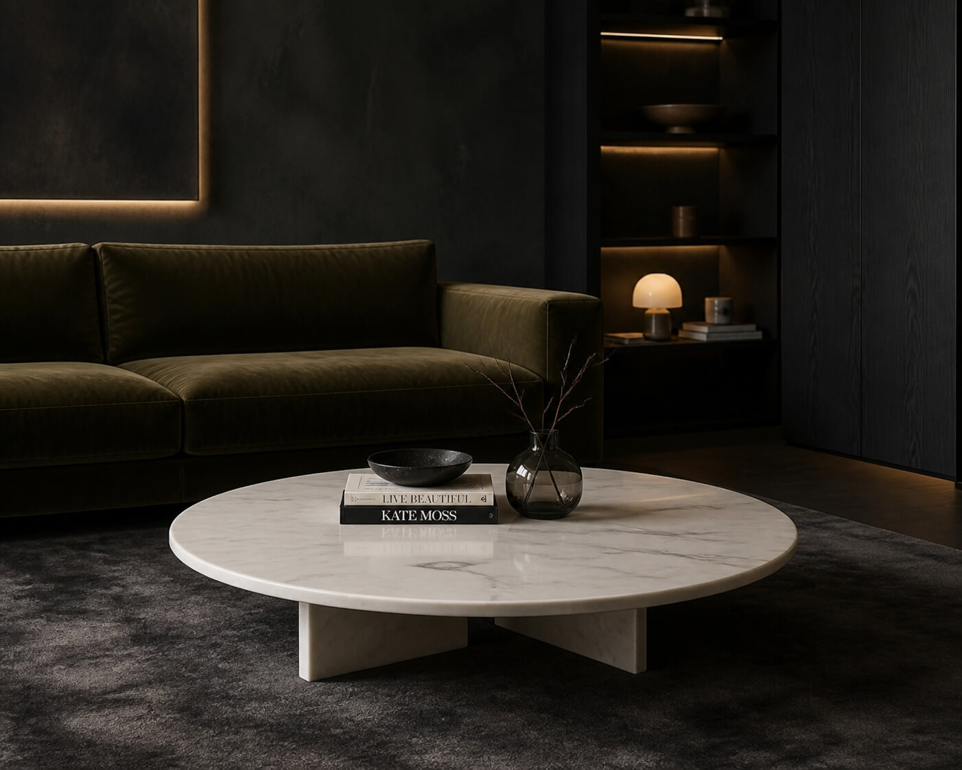 Round white marble coffee table in a dark moody lounge with olive velvet sofa, charcoal rug, and backlit display shelving.