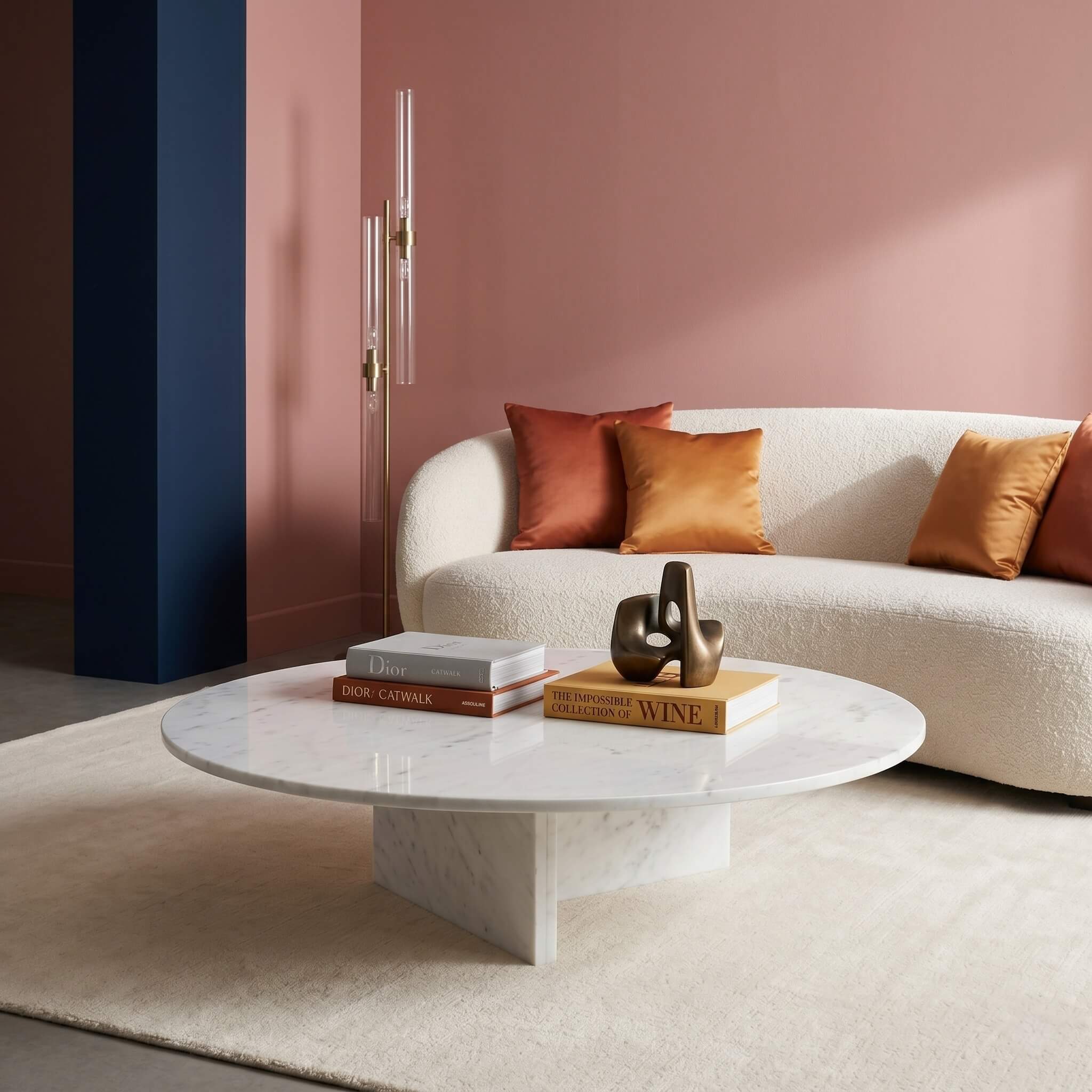 Round white marble coffee table on twin slab legs in glam living room, cream bouclé sofa, blush pink walls, brass lamp, topped with art books and bronze decorative sculpture.