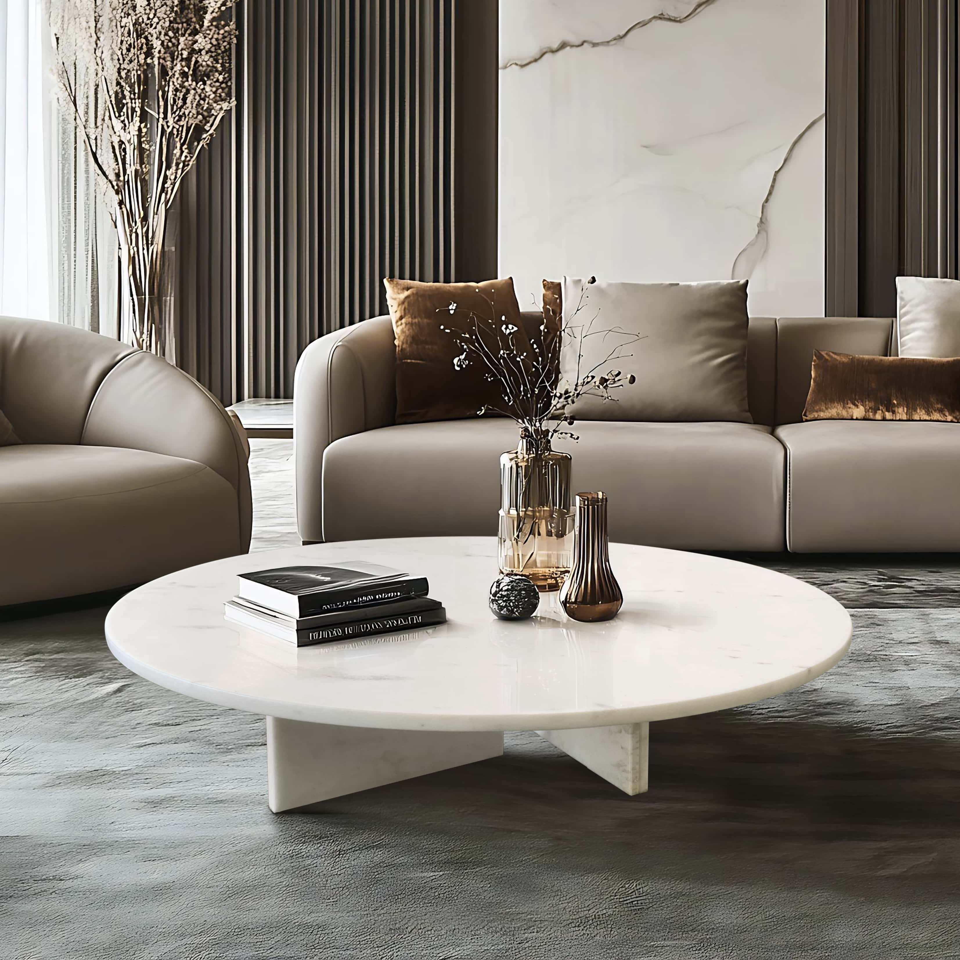 Round white honed marble coffee table against dark textured flooring in luxury interior, paired with grey leather sofa, marble accent wall, and amber glass vase styling.