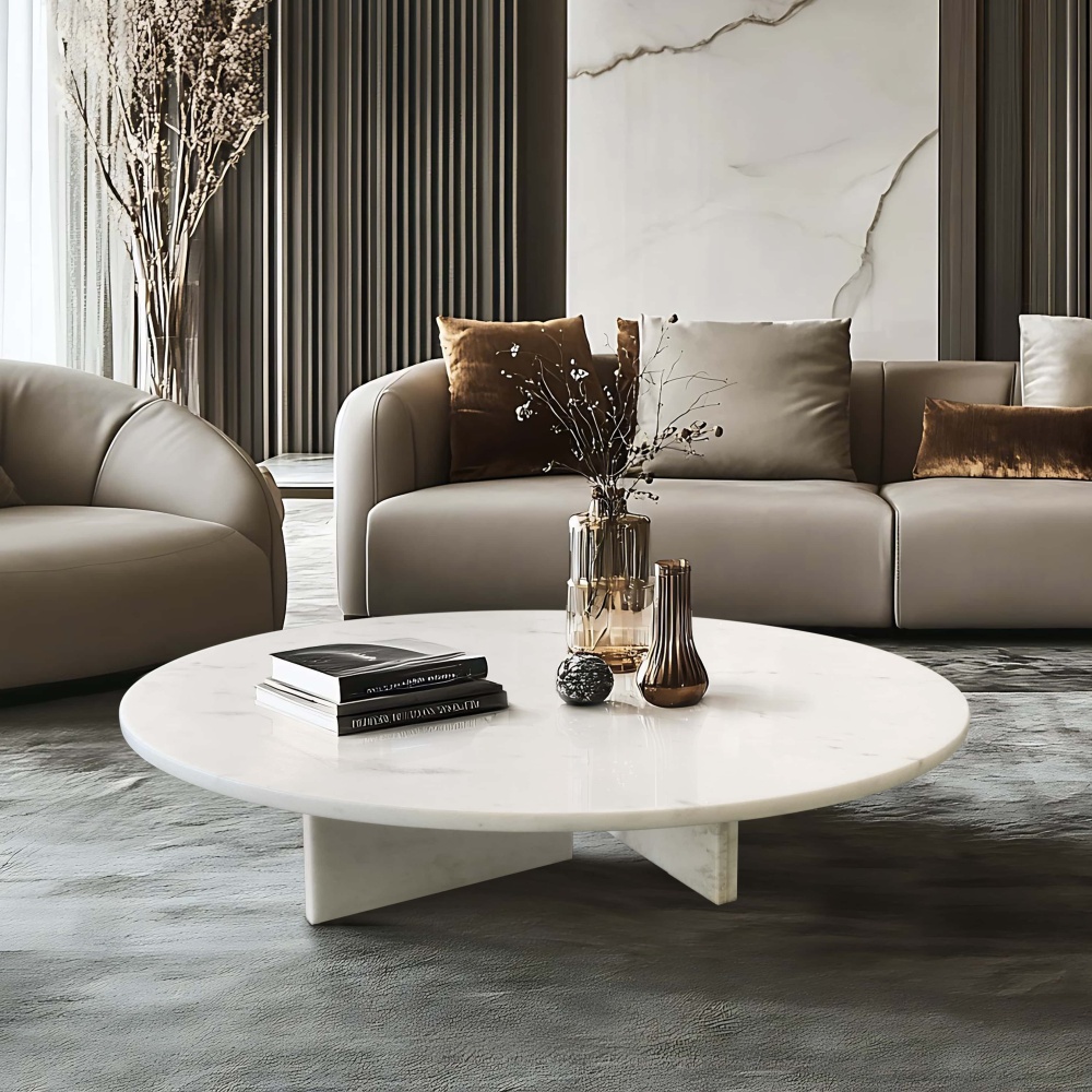 Round white honed marble coffee table against dark textured flooring in luxury interior, paired with grey leather sofa, marble accent wall, and amber glass vase styling.