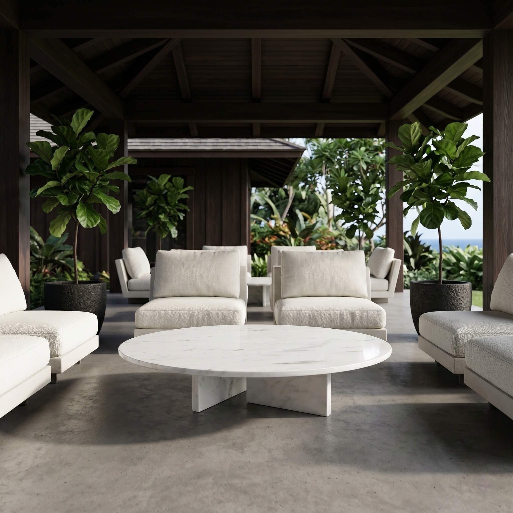 Round white marble coffee table on a covered tropical lanai with cream lounge chairs, fiddle-leaf figs, and concrete floor.