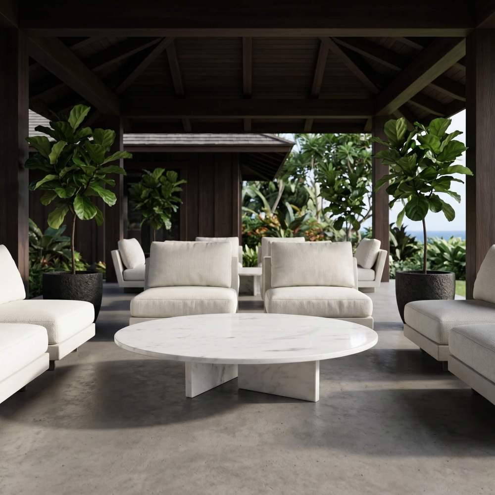 Round white marble coffee table on a covered tropical lanai with cream lounge chairs, fiddle-leaf figs, and concrete floor.