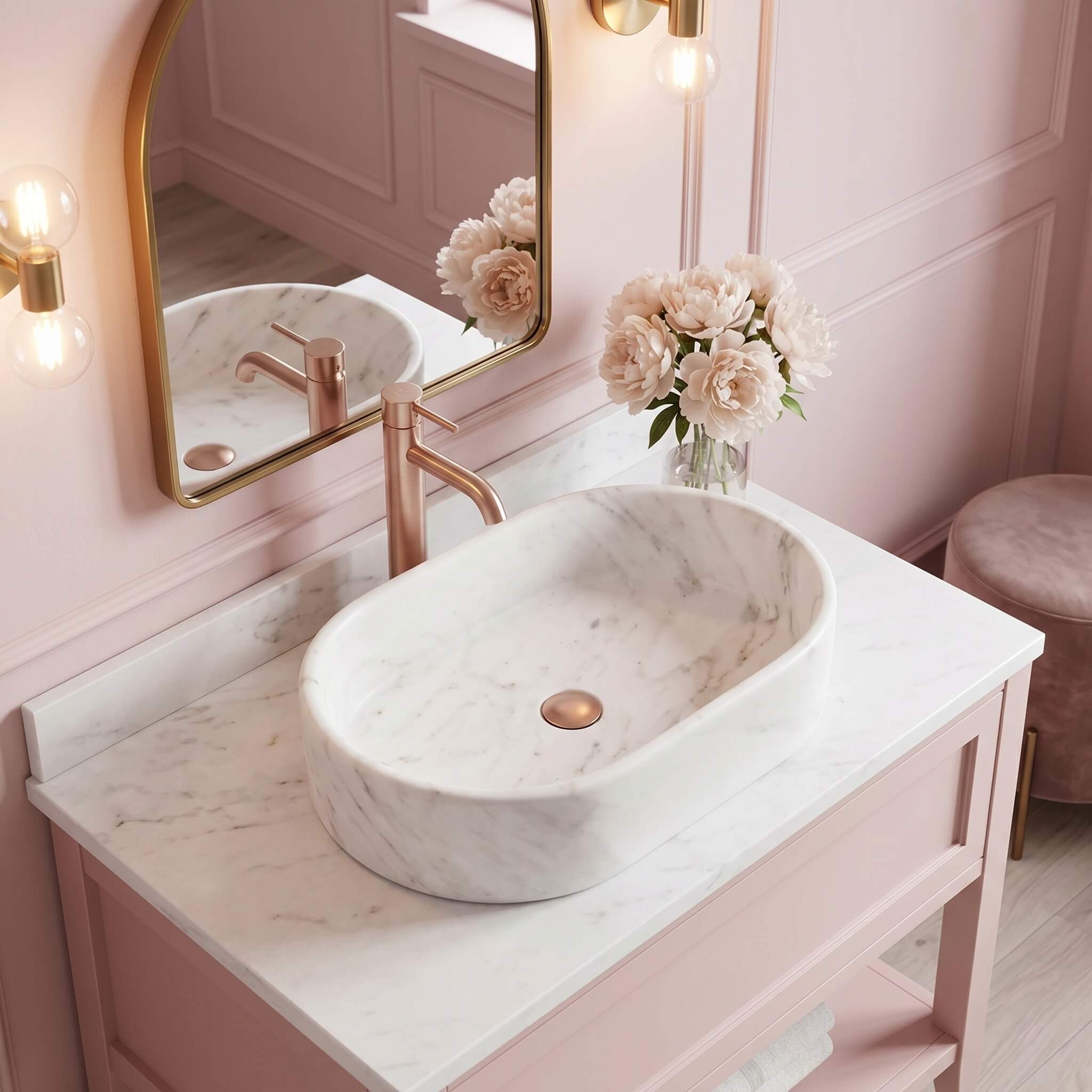 A Snow Mountain White marble oval vessel sink on a blush pink vanity with arched gold mirror, rose gold faucet and peonies in a romantic feminine glam bathroom.
