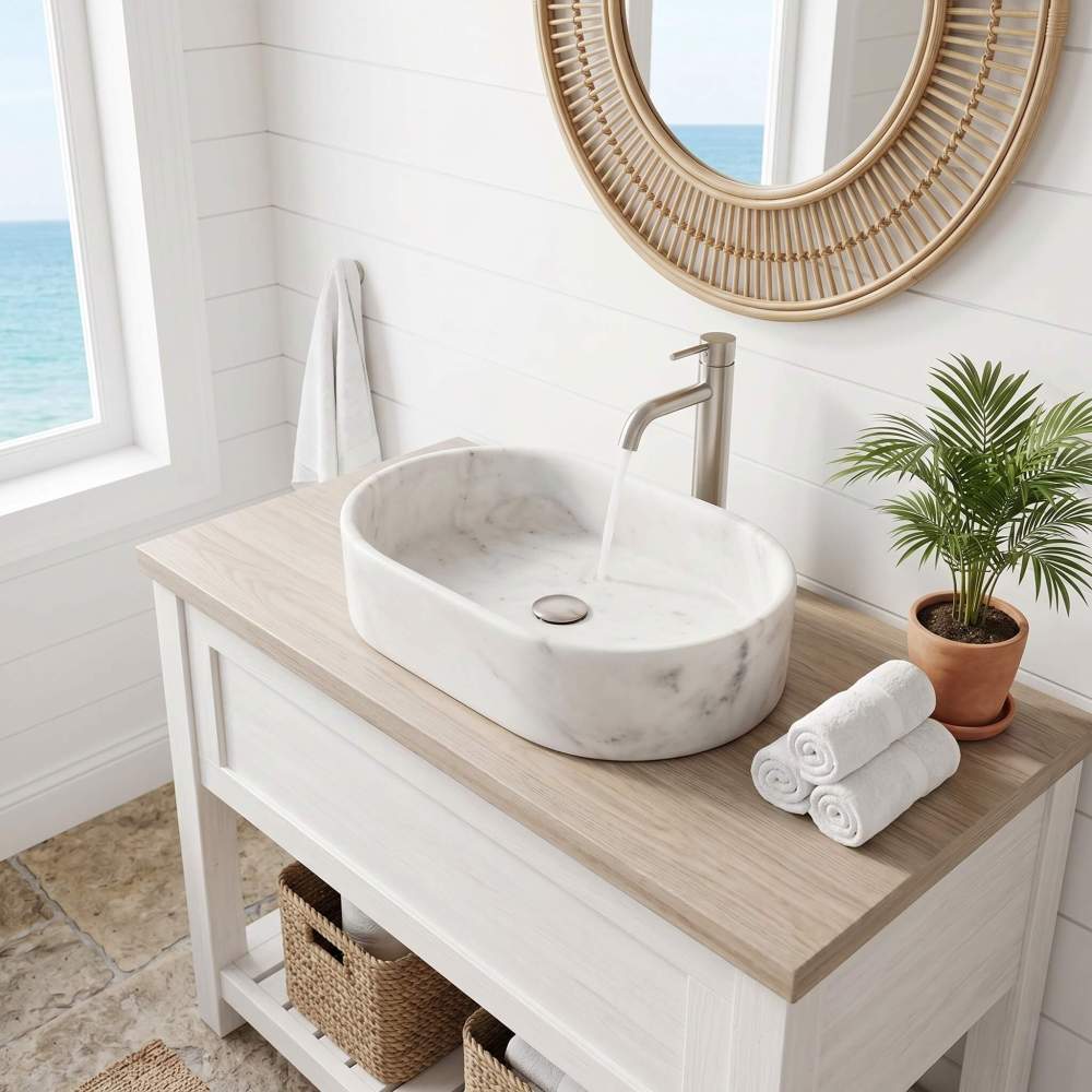 A Snow Mountain White marble oval vessel sink with running water on a light wood console against shiplap by an ocean-view window in a breezy coastal bathroom.