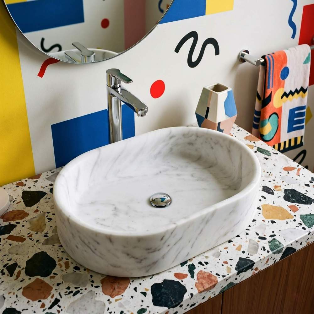 A Snow Mountain White marble oval vessel sink on a terrazzo top against bold geometric wall art with a chrome faucet in a playful Memphis postmodern bathroom.