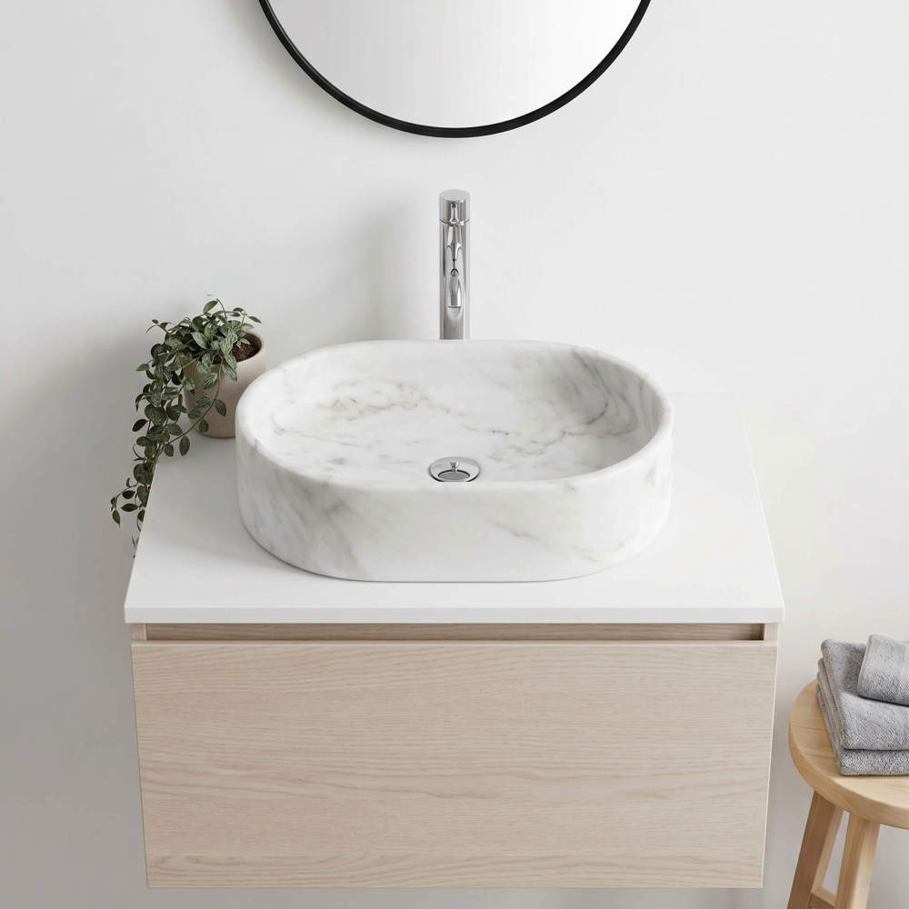 A Snow Mountain White marble oval vessel sink on a pale oak floating cabinet with a black round mirror, chrome faucet and trailing plant in a Nordic minimalist bath.