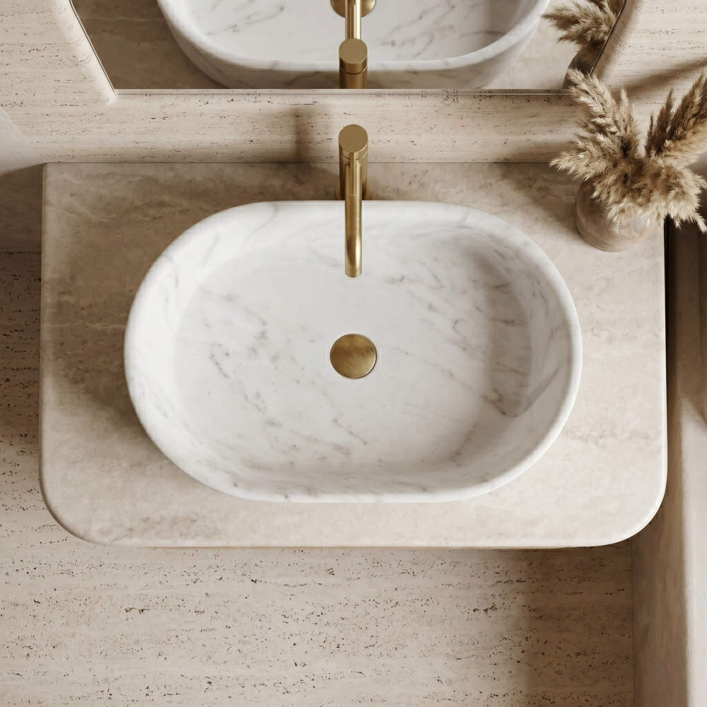 Overhead view of a Snow Mountain White marble oval vessel sink set into a travertine ledge with a gold faucet and pampas grass in a warm organic bathroom.