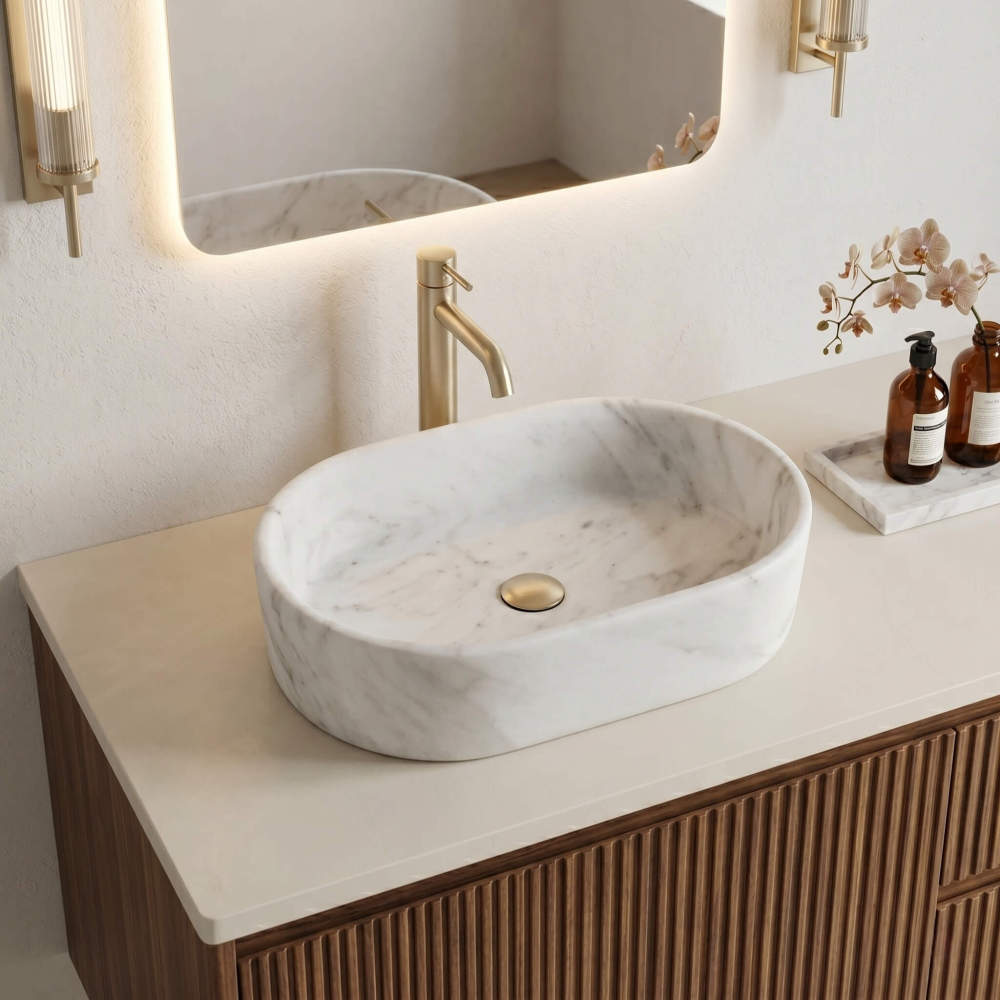 A single Snow Mountain White marble oval vessel sink on a fluted walnut vanity with backlit mirror, gold faucet and orchids in a warm modern luxe bathroom.