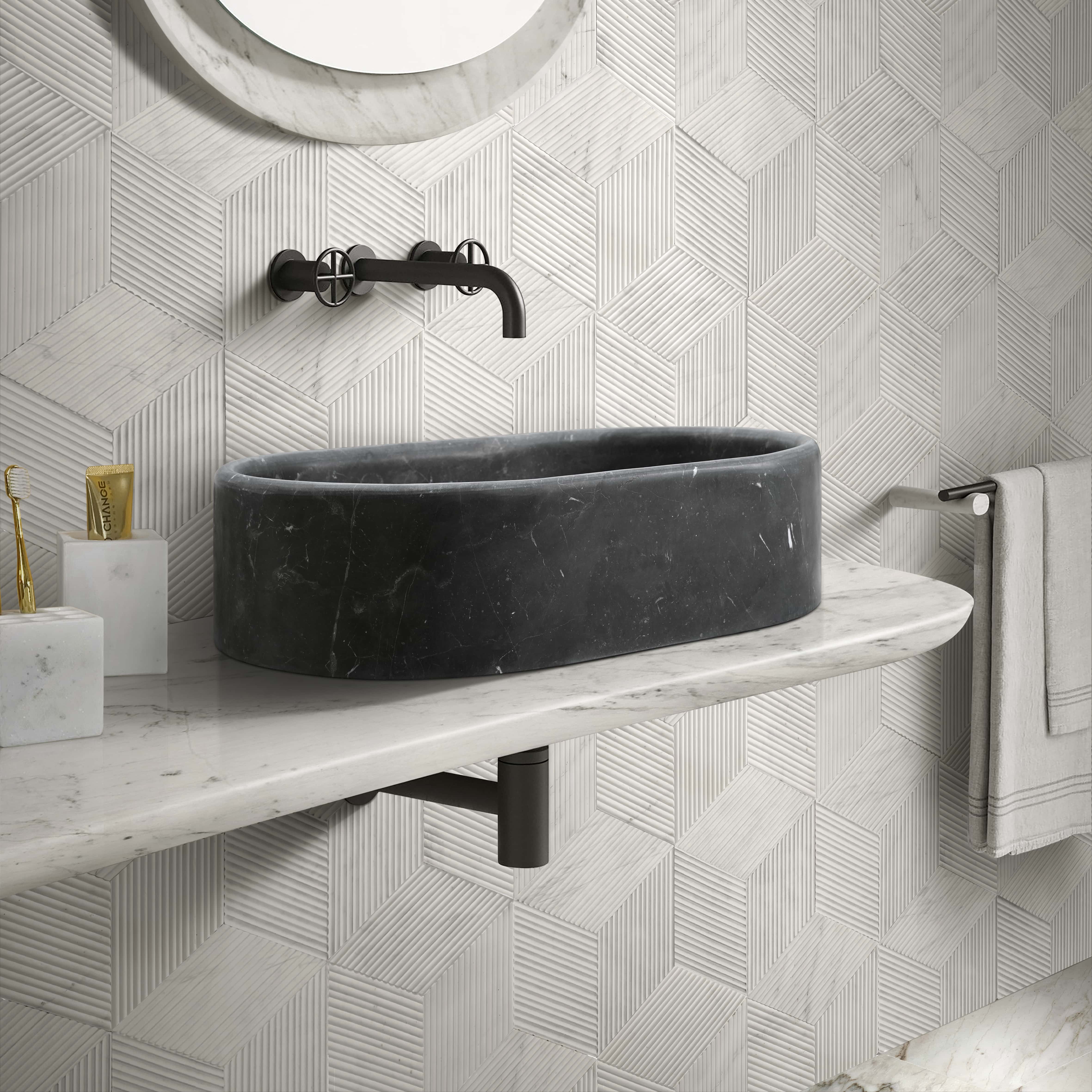 An Olive Black marble oval vessel sink against a Snow Mountain White hexagon textured wall tile, styled with honed white stone accessories.