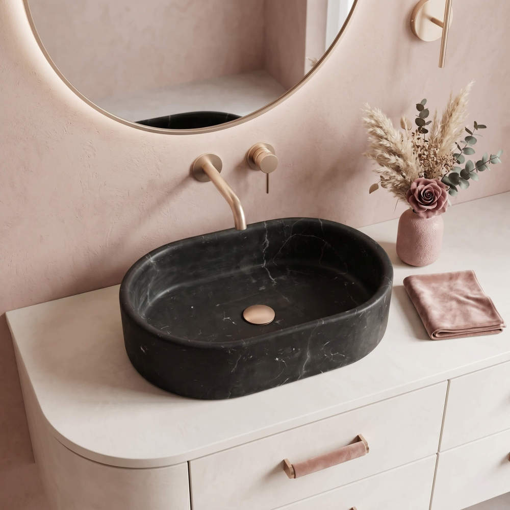 An Olive Black marble oval vessel sink on a cream curved vanity with a blush pink wall and champagne gold wall-mount faucet.