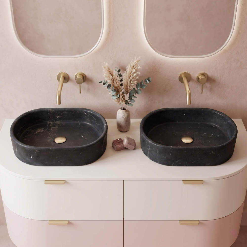 Two Olive Black marble oval vessel sinks on a blush pink curved vanity with gold wall-mount taps and oval backlit mirrors.