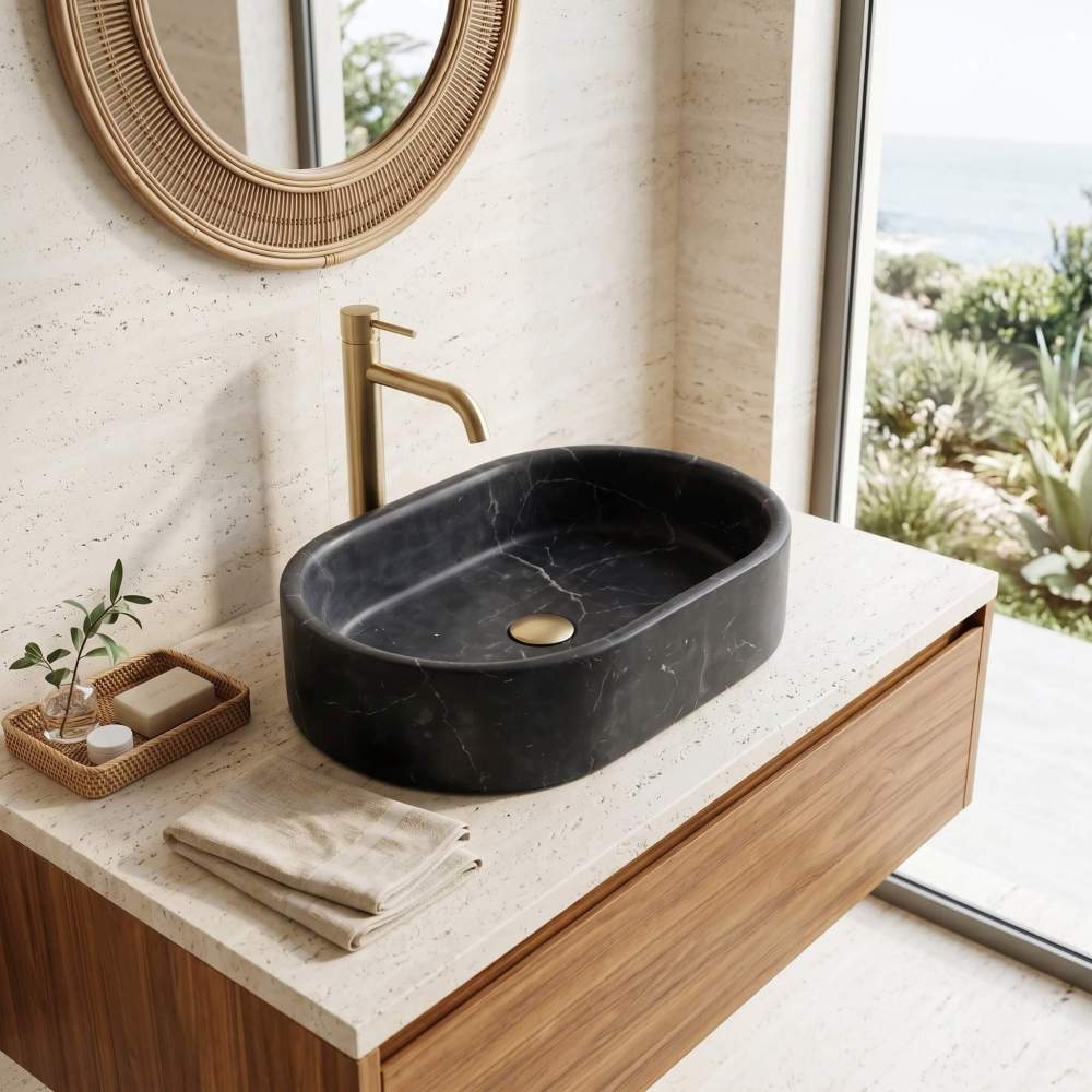 An Olive Black marble oval vessel sink on a travertine counter in a coastal bathroom with a rattan mirror and ocean window.