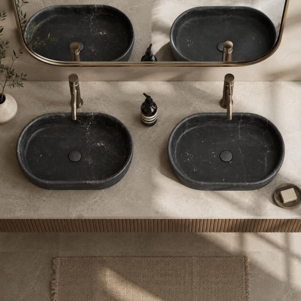 Overhead view of two Olive Black marble oval vessel sinks on a cream stone vanity with fluted wood base and bronze faucets.