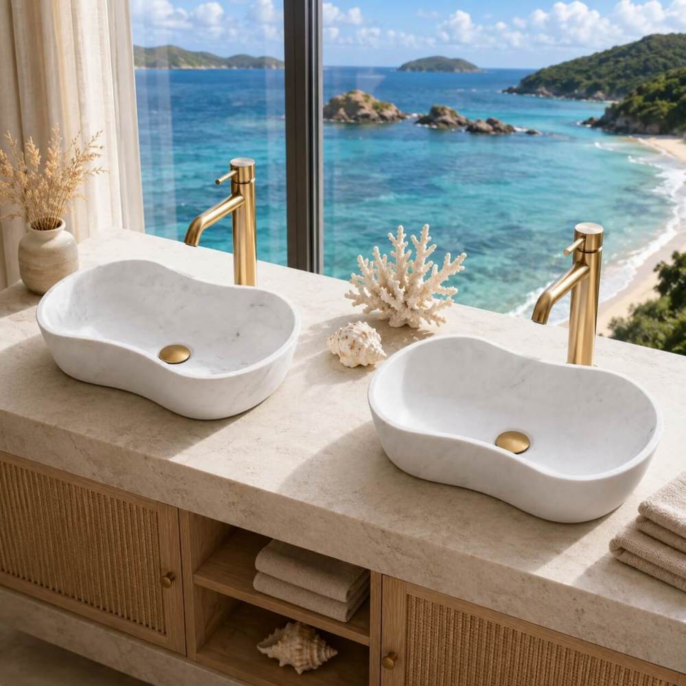Two Snow Mountain White honed wavy marble vessel sinks installed on a beige travertine countertop in a coastal resort style master bathroom with panoramic ocean views and brushed gold faucets.