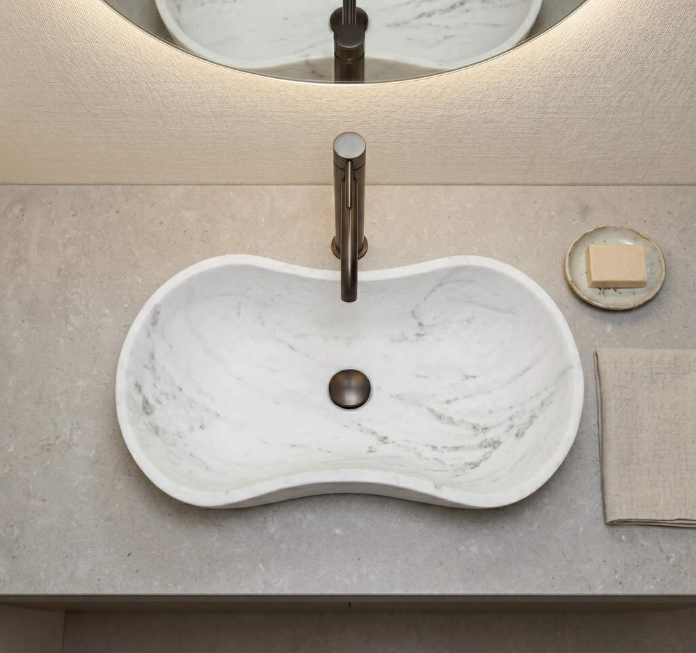 High-angle top-down perspective of the marble sink on a textured grey terrazzo stone countertop. Features a modern dark metal faucet and drain stopper, ceramic soap dish, and folded linen towel. Contemporary texture pairing.