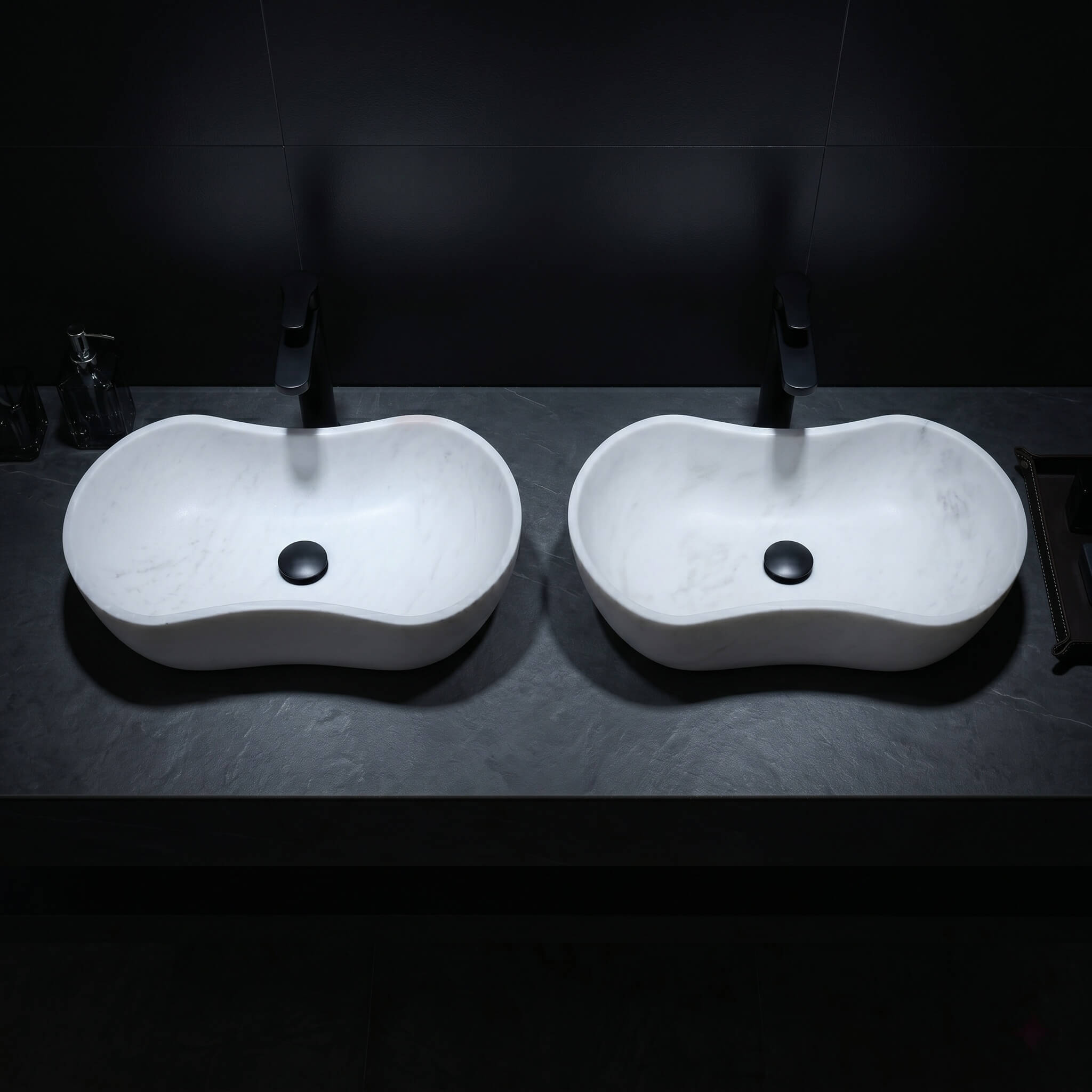 Top-down oblique shot of twin Snow Mountain White wavy marble basins on a textured dark charcoal slate countertop with matte black fixtures under dramatic minimalist spotlighting.