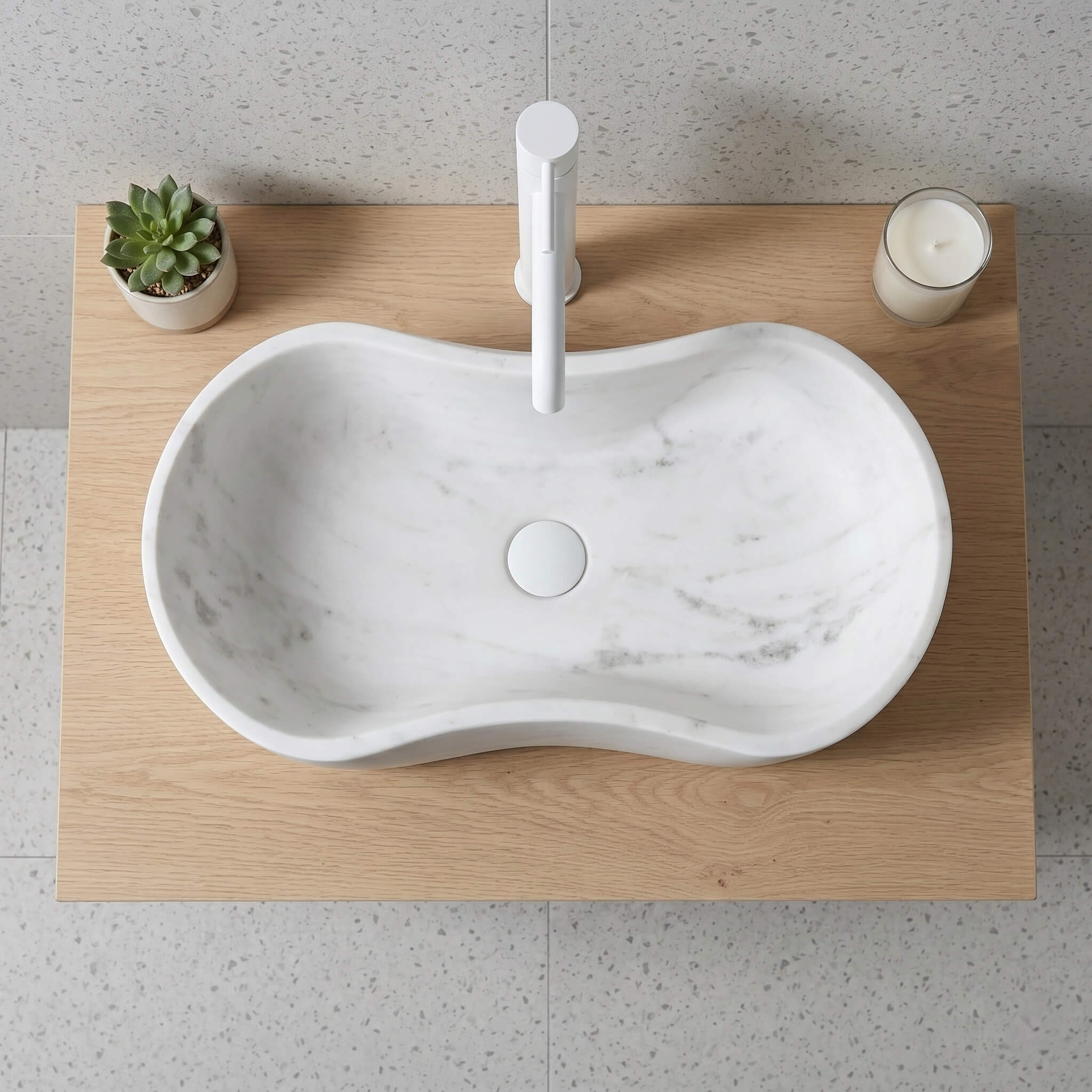 Direct overhead top-down view of the wavy marble sink centered on a light-wood countertop. Features a white minimalist faucet and drain, a potted succulent, and a candle in a glass. Contemporary and clean.