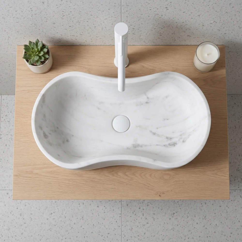 Direct overhead top-down view of the wavy marble sink centered on a light-wood countertop. Features a white minimalist faucet and drain, a potted succulent, and a candle in a glass. Contemporary and clean.