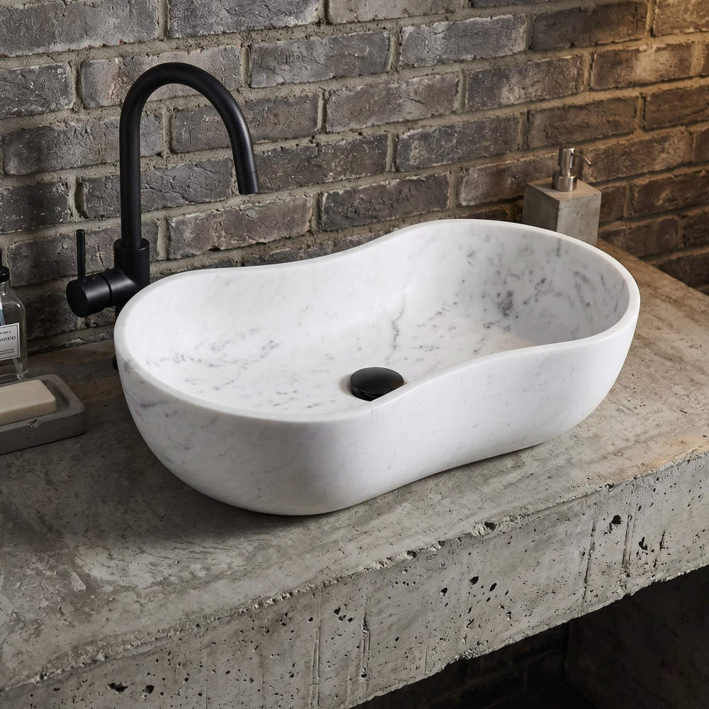 Front-on, eye-level perspective of the wavy marble vessel sink on a rough concrete countertop against an exposed brick wall. Combined with an industrial matte black gooseneck faucet and drain. Industrial urban chic.