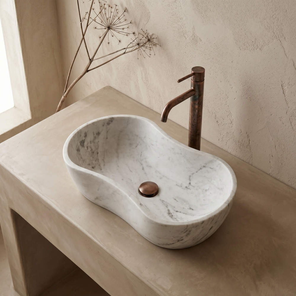 Eye-level shot of the marble vessel sink with wavy rim on a beige concrete vanity against textured walls. Matched with an aged copper gooseneck faucet and drain, and a dried botanical accent. Rustic and natural feel.
