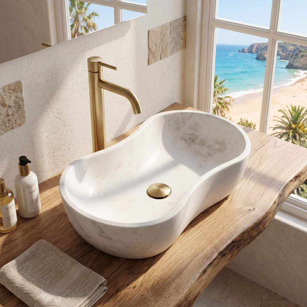 High-angle perspective of the marble sink on a live-edge wood plank vanity by a large window overlooking a coastal beach and ocean view. Polished gold faucet and drain, and toiletries. Luxury resort design.