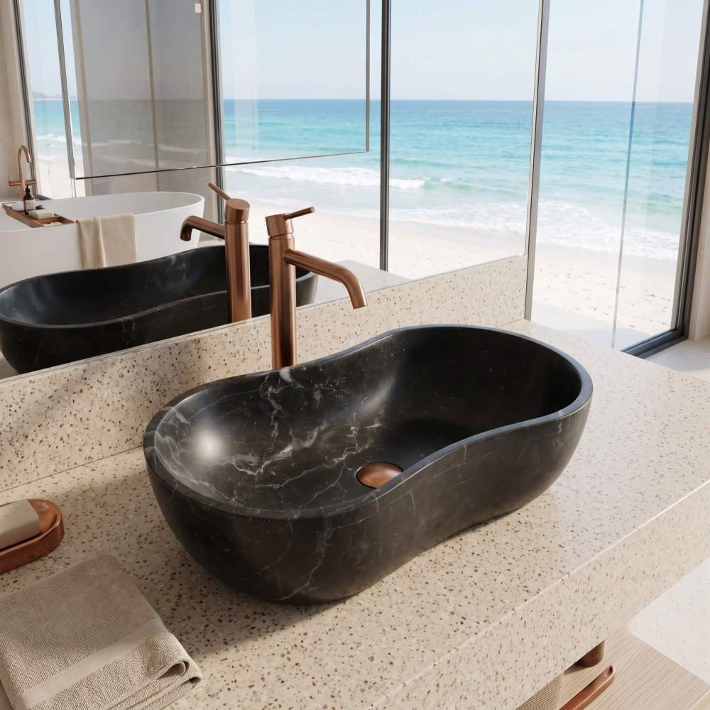 A honed Olive Black natural marble wave vessel sink on a speckled terrazzo vanity, framed by an expansive coastal ocean beachfront view.