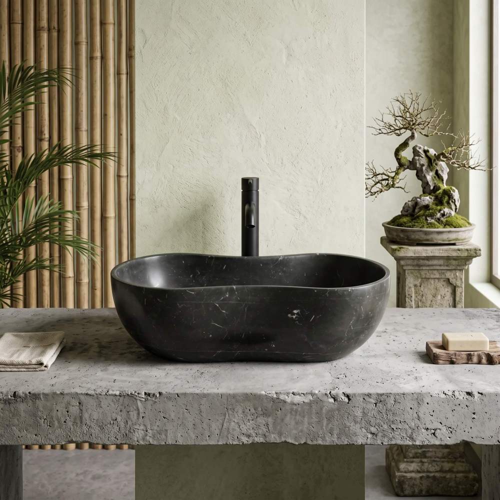 Front view of a honed Olive Black marble wave vessel sink on a raw concrete surface, surrounded by minimalist wabi-sabi elements and bamboo.