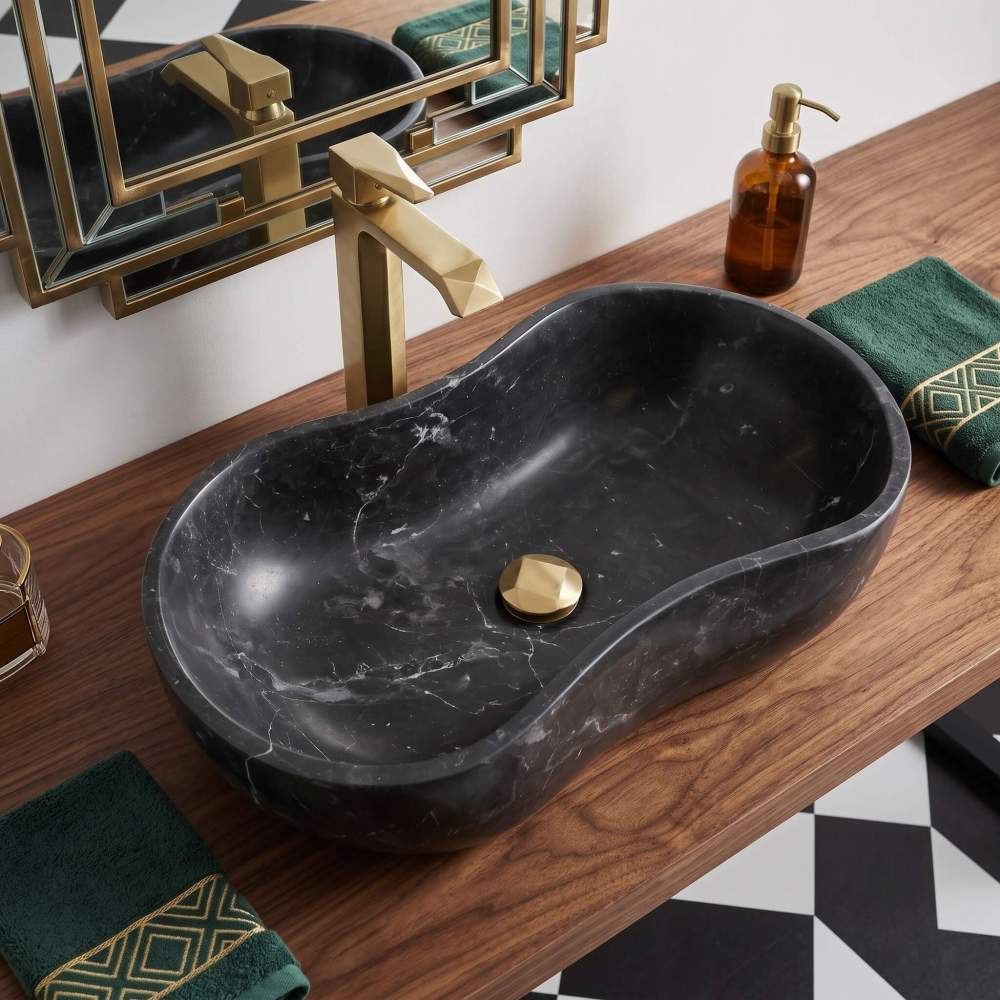 An elegant Olive Black marble wave shape stone sink on a walnut vanity top, complemented by a geometric brass faucet and checkered flooring.