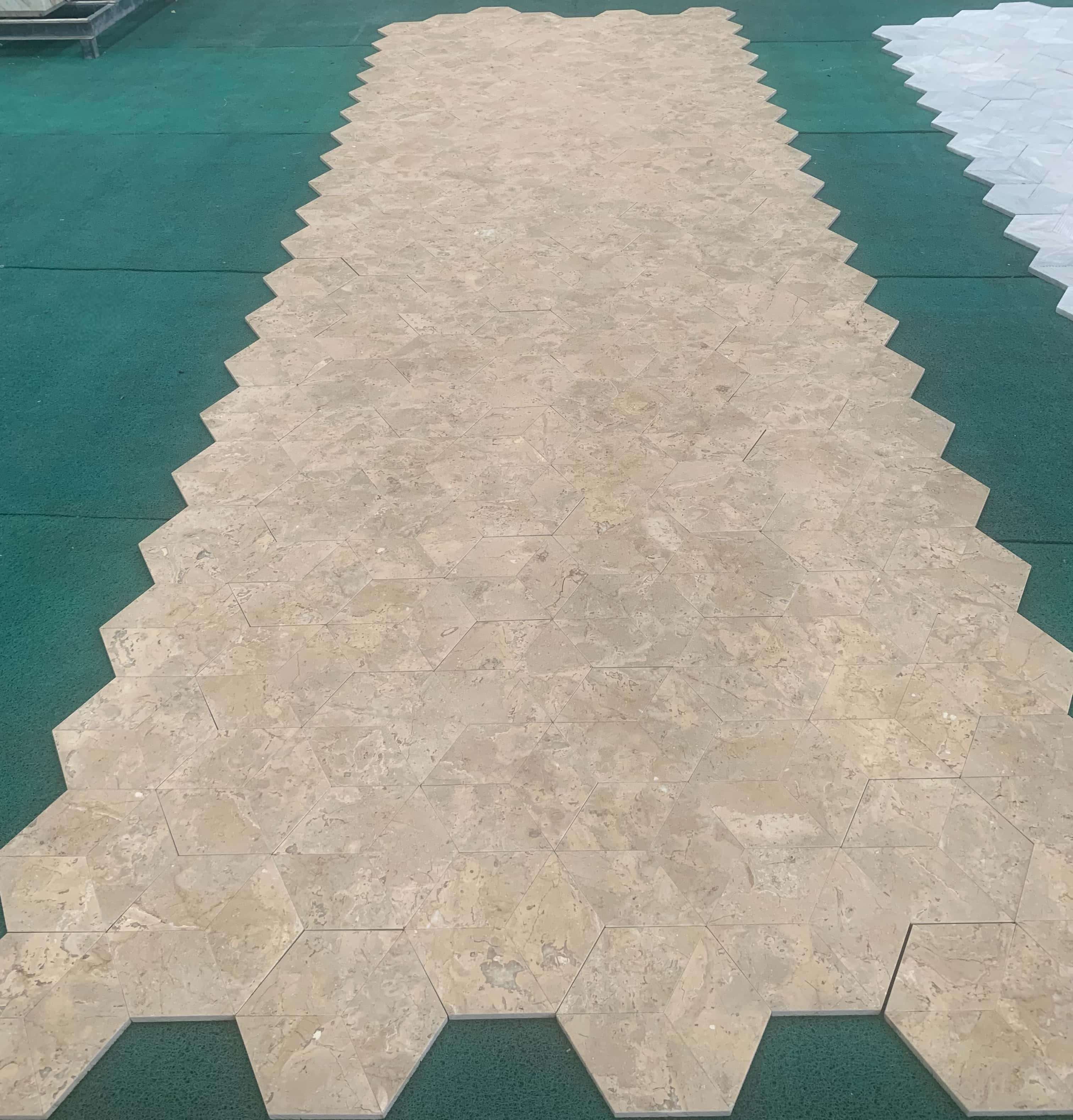 Factory dry-lay inspection of Honed Amber Gold marble 3D cube hexagon tiles on green surface for tone matching.