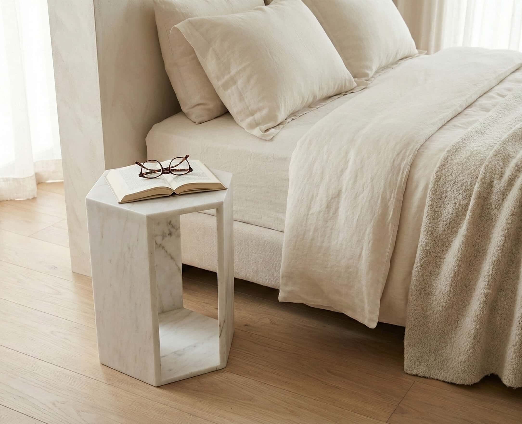 A honed Snow Mountain White marble hexagon side table used as a nightstand beside a beige linen bed holding an open book.