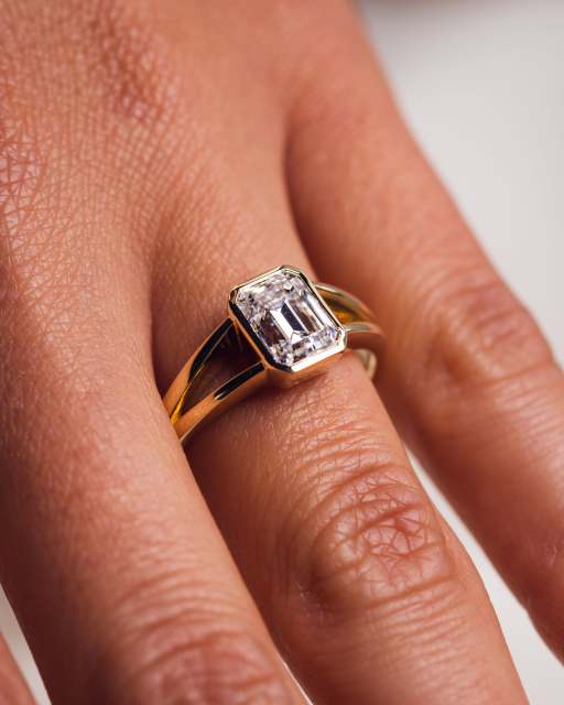 Angled view on a hand showing the split shank band and bezel setting of an emerald cut lab diamond ring in 14K yellow gold.