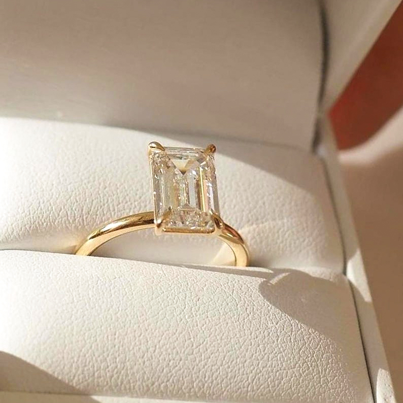 A stunning 4-carat emerald cut lab-grown diamond ring presented in a luxury white jewelry box under warm lighting, proving its No BGM crystal transparency.