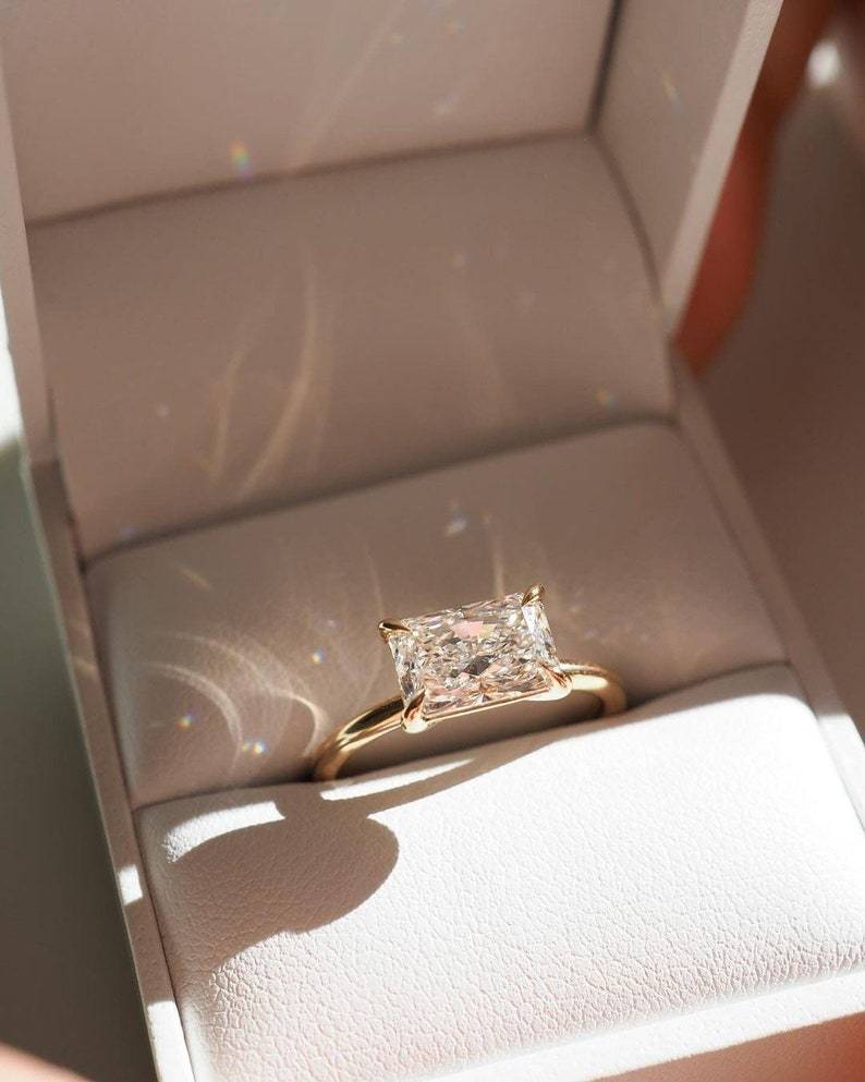 Top-down perspective of a 1.1 carat radiant cut lab grown diamond in a 14K solid gold east-west horizontal engagement ring setting, resting inside a premium presentation box and showcasing D color and VVS2 clarity brilliance under natural light.