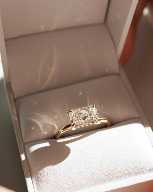 Top-down perspective of a 1.1 carat radiant cut lab grown diamond in a 14K solid gold east-west horizontal engagement ring setting, resting inside a premium presentation box and showcasing D color and VVS2 clarity brilliance under natural light.