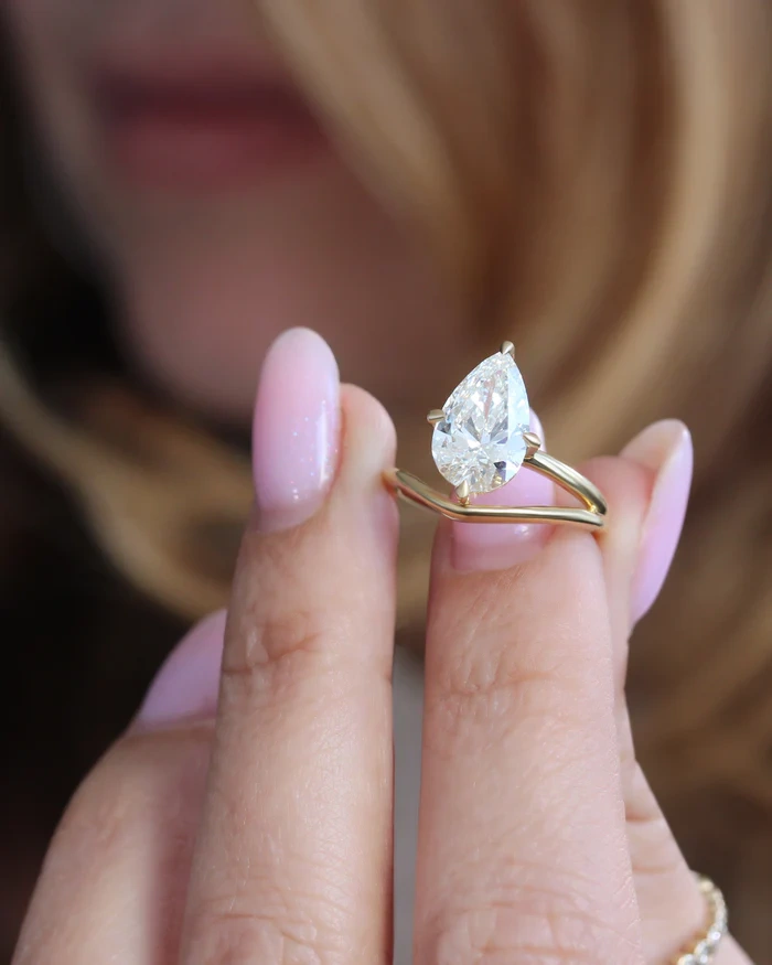 Close-up of a 3 carat pear lab grown diamond ring held between fingers, showing the intricate craftsmanship of the claw prongs and the brilliant E color stone.