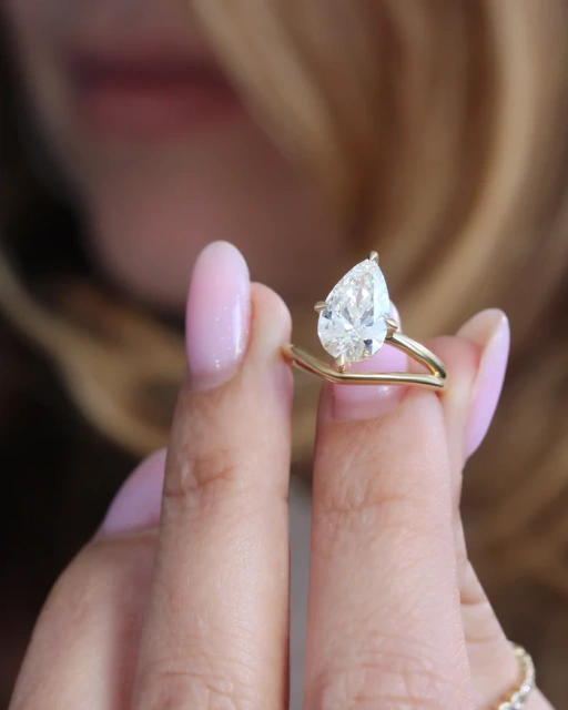 Close-up of a 3 carat pear lab grown diamond ring held between fingers, showing the intricate craftsmanship of the claw prongs and the brilliant E color stone.